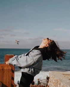 a woman standing next to the ocean with her hair blowing in the wind and birds flying overhead