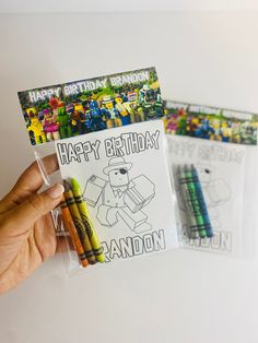 a hand holding up a birthday card with crayons in front of it and a coloring book next to it