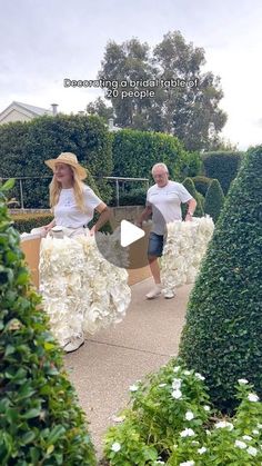 two people are walking through a garden with flowers in the center and bushes behind them