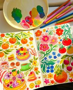 an open coloring book on a table next to colored pencils and a bowl of fruit