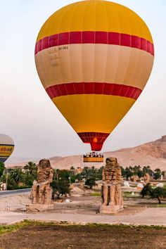 two hot air balloons flying in the sky over some stone statues and palm trees with mountains in the background