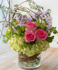 a glass vase filled with pink roses and greenery on top of a wooden table