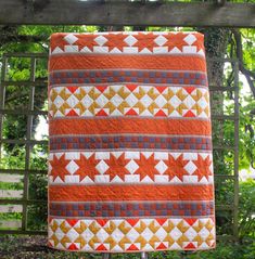 an orange and white quilt hanging on a wooden fence with trees in the back ground