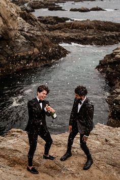 two men in tuxedos are standing on rocks near the ocean and one is holding a bottle