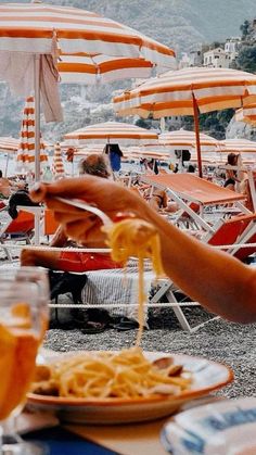 an outdoor dining area with orange umbrellas and people eating spaghetti on the beach in front of them