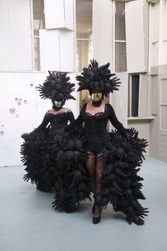 two women dressed in black are standing next to each other with large feathers on their head