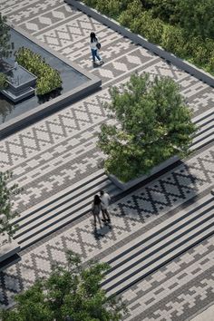 two people are walking down the sidewalk in front of some trees and benches with their shadows on them