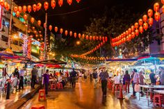 many people are walking through an outdoor market area at night with red lanterns hanging above them