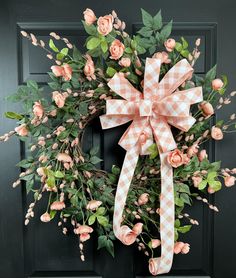 a wreath with pink flowers and green leaves hanging on a black front door, decorated with gingham ribbon