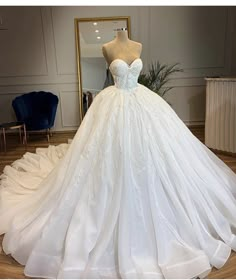 a white wedding dress on display in a room with a mirror and blue chair behind it