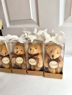 four teddy bears in small clear boxes with white ribbons and name tags on them, sitting next to each other