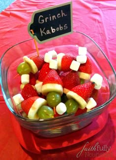 a glass bowl filled with fruit topped with marshmallows and apples next to a sign that says grinch kabobs