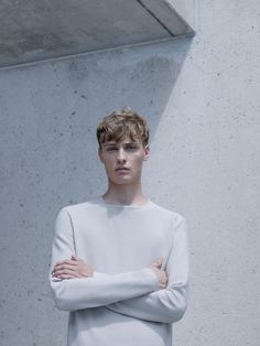 a young man with his arms crossed standing in front of a concrete wall wearing a white sweater