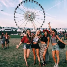 three girls standing in front of a ferris wheel at an outdoor music festival with their arms up