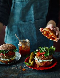 a person in an apron holding a sandwich over two plates with pickles and tomatoes on them