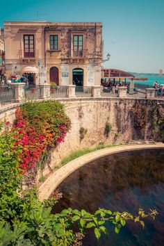 people are sitting on the side of an old building overlooking water and flowers in front of it