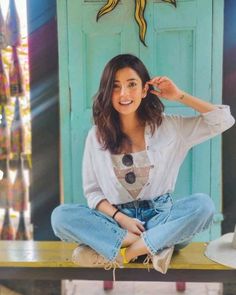 a woman sitting on top of a wooden bench in front of a blue door wearing jeans and a white shirt