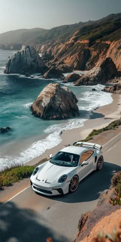 a white sports car driving down the road next to the ocean and rocky coastline with large rocks in the background