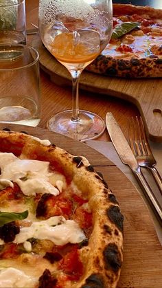 two pizzas sitting on top of a wooden table next to silverware and wine glasses