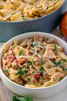 two bowls filled with pasta and meat on top of a wooden table next to an apple