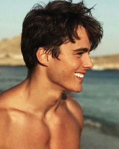 a man with no shirt on standing next to the ocean and smiling at something he's holding in his hand