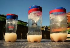 three plastic containers filled with liquid sitting on top of a cement ground next to a fence