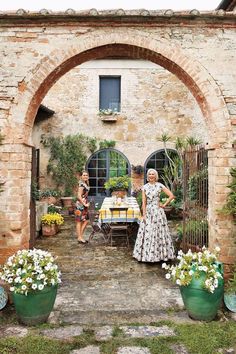 two women standing in front of a brick building with potted plants and flowers on the ground