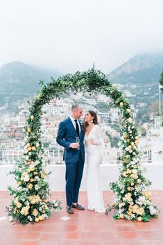 a bride and groom are standing under an arch with flowers on it, surrounded by greenery