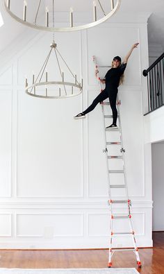 a woman standing on top of a ladder next to a chandelier in a white room