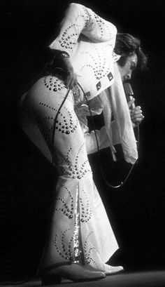 elvis presley performing on stage in white dress and black hat with his hands behind his back