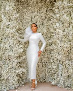 a woman standing in front of a wall with white flowers and greenery on it