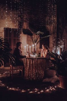 a man and woman sitting at a table with candles in front of them, surrounded by potted plants