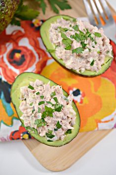 two avocados filled with tuna salad on top of a wooden cutting board next to a knife and fork