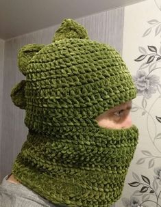 a person wearing a green knitted hat and neck gaiter with flowers on the wall behind them