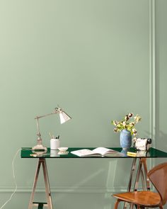 a desk with a lamp, books and flowers on it in front of a green wall