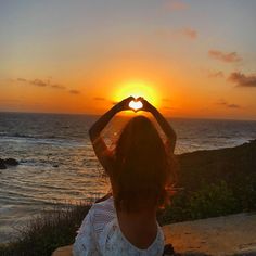 a woman sitting on the edge of a cliff with her hands in the shape of a heart