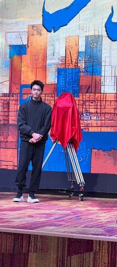 a man standing on top of a stage next to a red cloth covered chair and umbrella