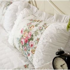 a white bed topped with lots of pillows next to an alarm clock on top of a night stand