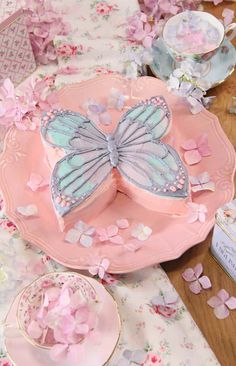 a pink plate topped with a butterfly shaped cake next to tea cups and saucers