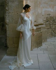 a woman standing in front of a stone wall wearing a white dress with sheer sleeves