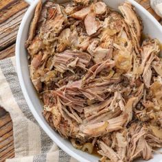 a white bowl filled with shredded meat on top of a wooden table next to a napkin