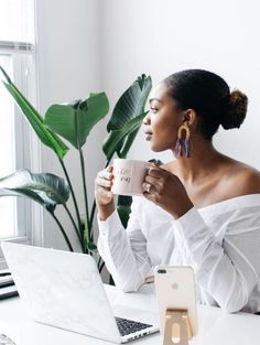 a woman sitting at a desk with a laptop and cup in her hand while holding a mug