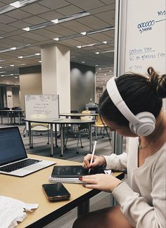 a woman sitting at a desk with headphones on, writing in her notebook and using a laptop