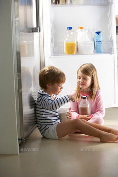 two children sitting on the floor in front of an open refrigerator with milk and juice