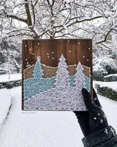 a hand holding up a piece of string art in the snow with trees on it