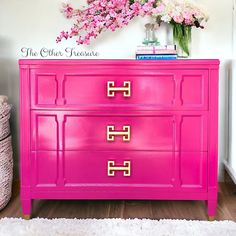 a bright pink dresser with gold handles and flowers on the top, in front of a white wall