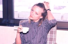 a woman sitting in a chair holding a coffee cup and looking at the camera with her hand on her head