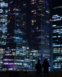 two people are standing in front of some tall buildings at night with lights on them