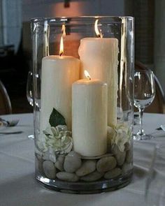 three white candles are sitting in a glass vase with rocks and greenery on the table