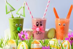 three glasses filled with different colored drinks sitting on top of grass next to eggs and flowers